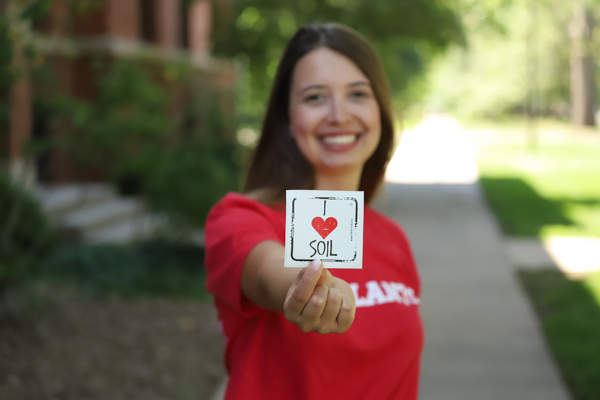 woman standing on sidewalk holding I Heart Soil sticker